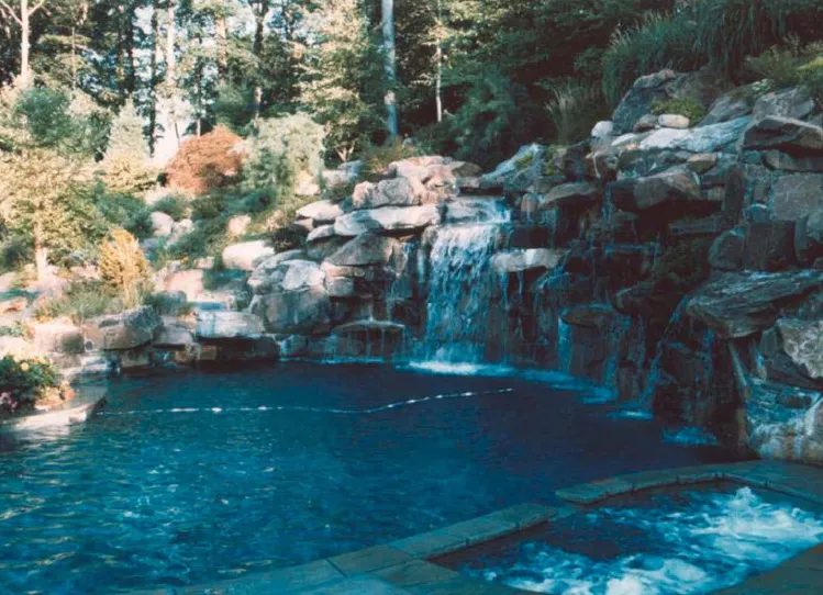 Freeform pool with a large natural boulder waterfall wall and wooded hillside backdrop in New Jersey by Gorlin Pools