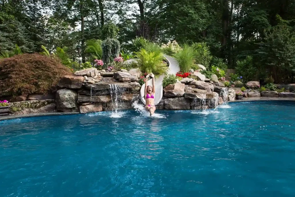 Child sliding into a freeform pool with rock waterfall feature and lush backyard landscaping in New Jersey by Gorlin Pools