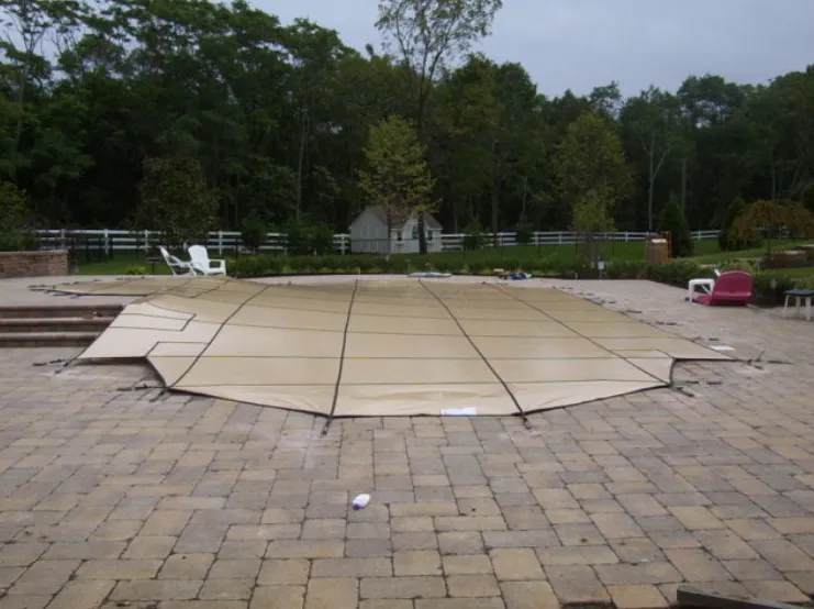 Freeform pool with tan mesh safety cover anchored to a brick paver patio surrounded by wooded backyard in Lakehurst, NJ by Gorlin Pools