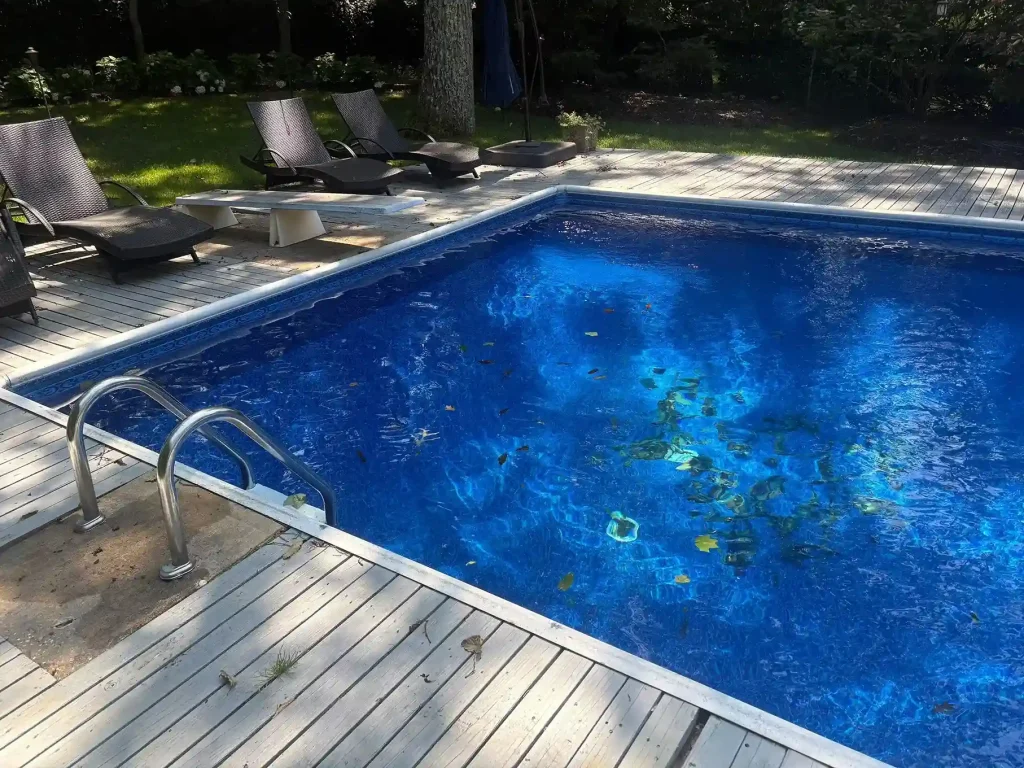 Rectangular pool with leaves and debris floating in water, stainless steel handrail, and wood deck with lounge chairs in New Jersey by Gorlin Pools